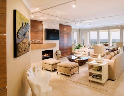 宽敞的米色客厅，铺着瓷砖地板，墙壁上有木板，有沙发、椅子和软凳，一个可以看到大海的阳台，一个小壁炉，壁炉上方有一台电视