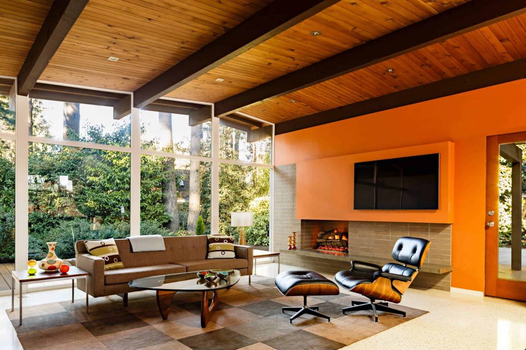 房子里有宽敞的客厅，有窗墙，木质天花板，棕色沙发和黑色扶手椅，棕色砖壁炉，壁炉上方橙色墙壁上有一台电视