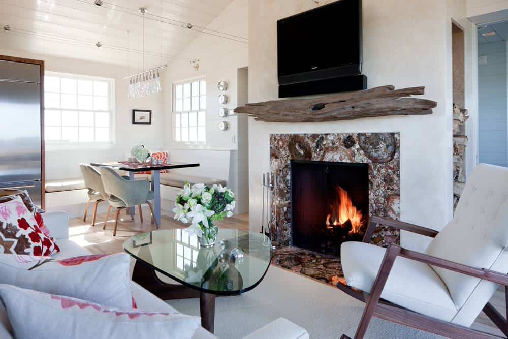 海滩风格的小客厅紧挨着厨房，房子里有白墙、沙发和摇椅、棕色花岗岩壁炉、电视和壁炉上方的一根原木