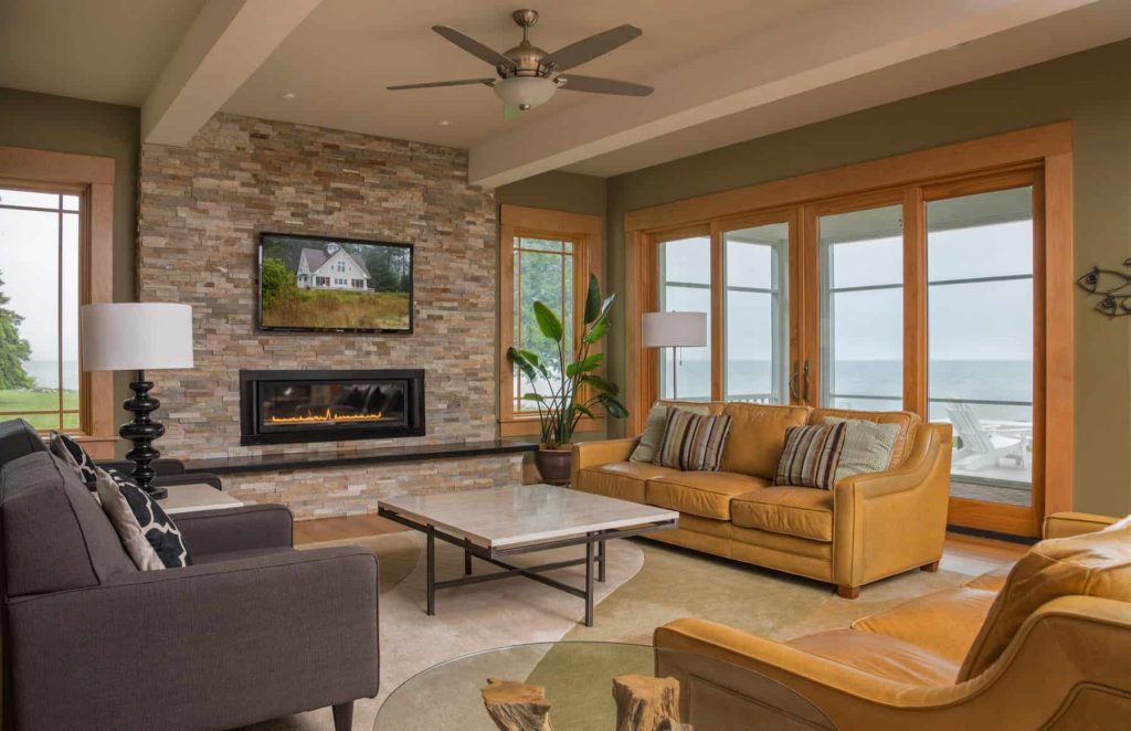 房子里有一个中等大小的客厅，可以看到海景，绿色的墙壁，棕色和灰色的沙发，石瓦墙上的小壁炉，壁炉上方有一台电视